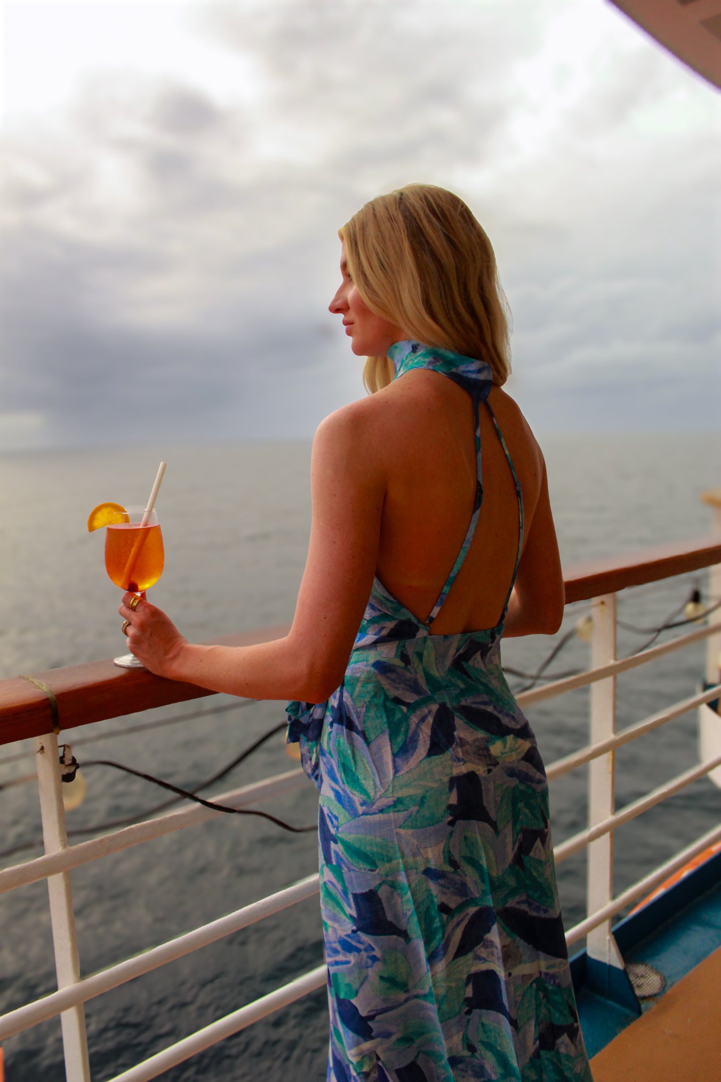 Woman in a blue tropical print halter dress enjoying a drink on a cruise ship balcony.