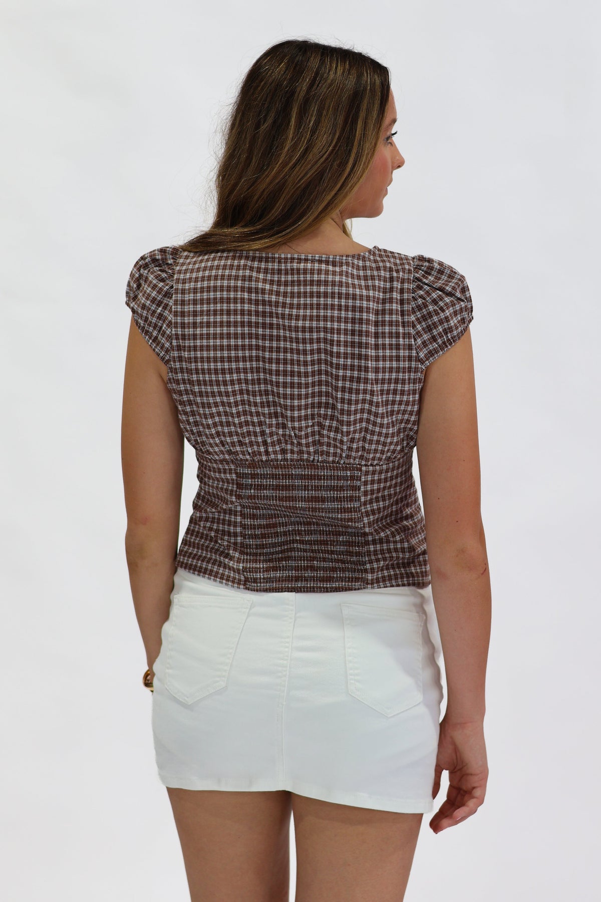 A woman shown from the back wearing a brown and white gingham check short-sleeve top with puff sleeves and a gathered waist, paired with white shorts, against a plain white background.