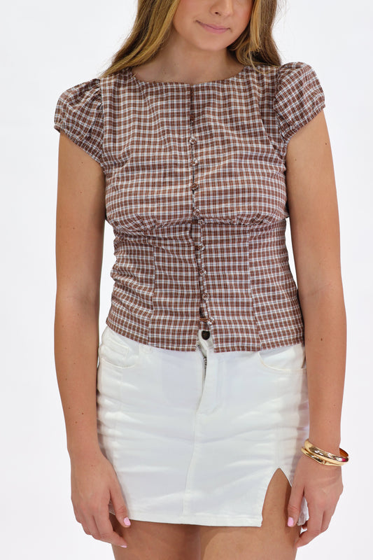 A woman wearing a brown and white gingham check top with puff sleeves and a gathered front, paired with a white mini skirt, against a plain white background.