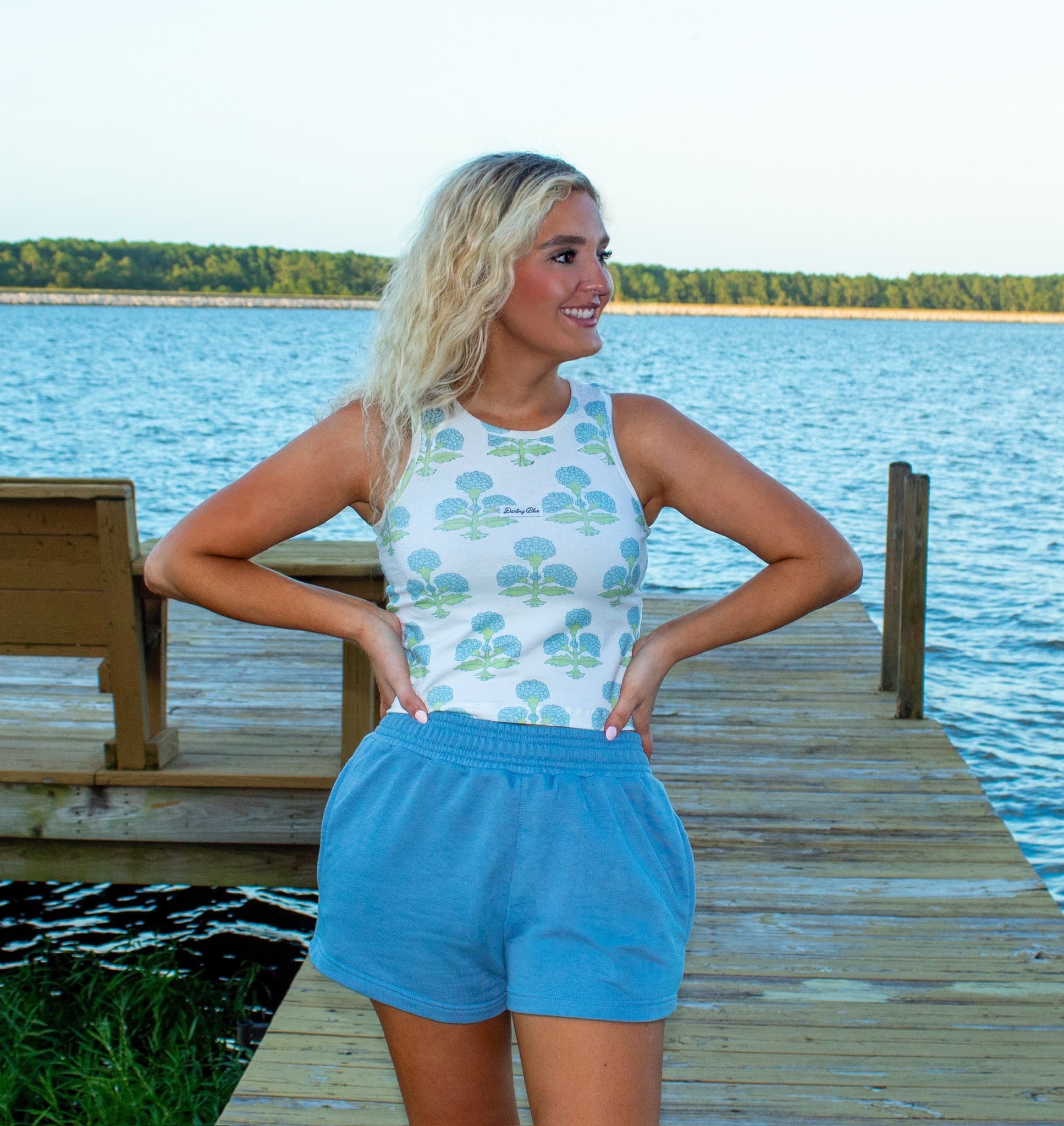 Woman standing on a dock by a lake wearing a sleeveless top and shorts.