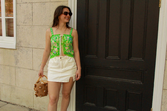 Woman wearing a green embroidered crop top and white mini skirt, accessorized with sunglasses and a handbag.