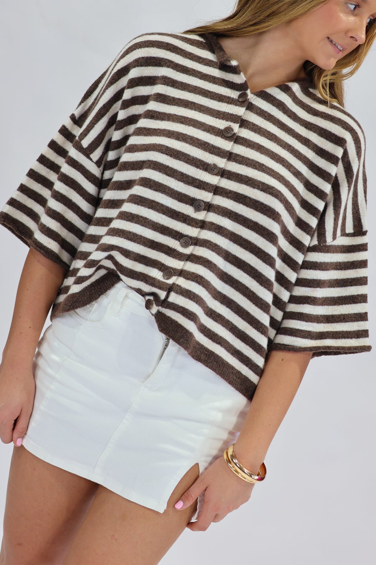 A woman wearing a brown and cream striped button-up knit top with wide three-quarter sleeves and a white mini skirt, photographed against a light gray studio background.