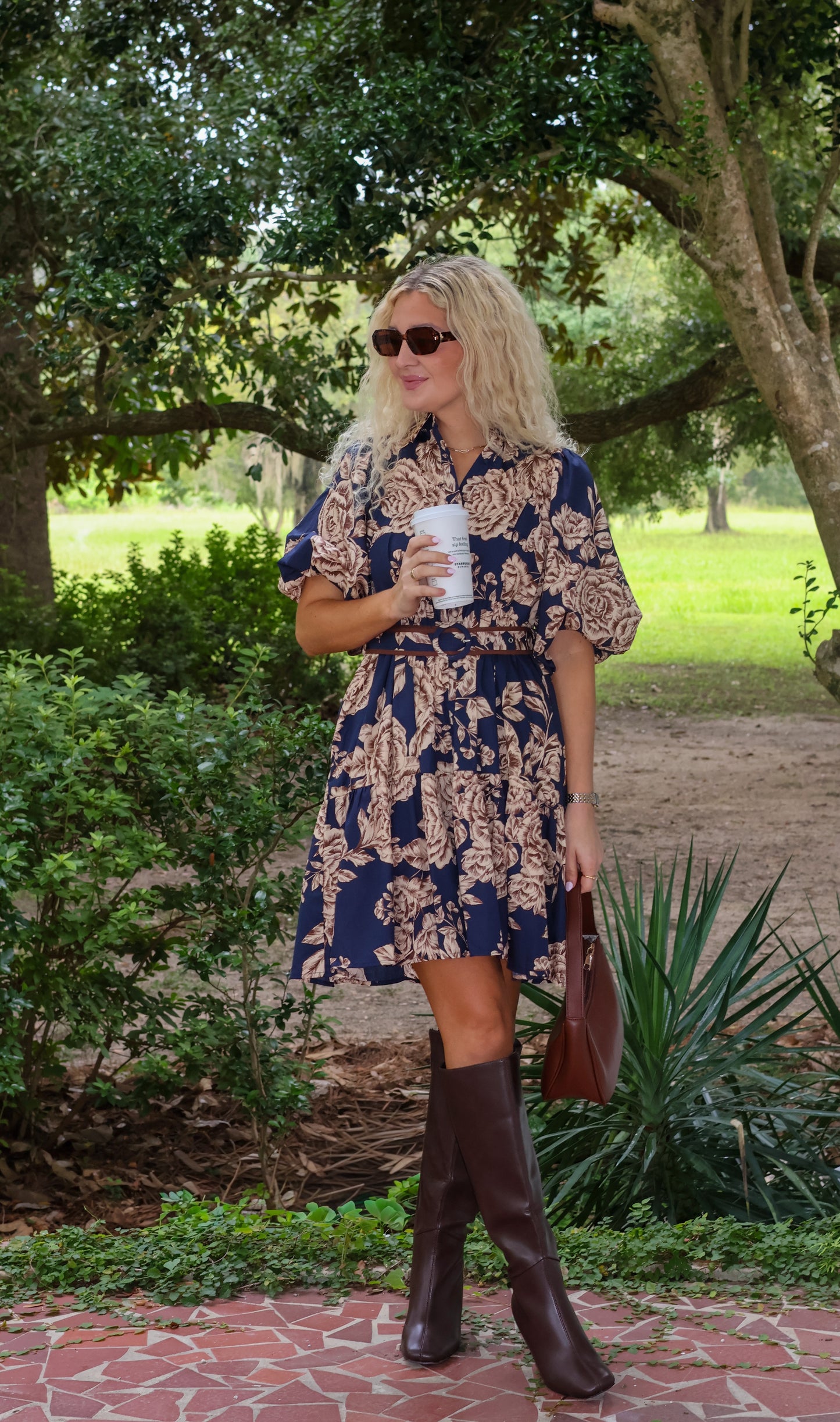 Woman in a floral dress and brown boots holding a coffee cup outdoors.