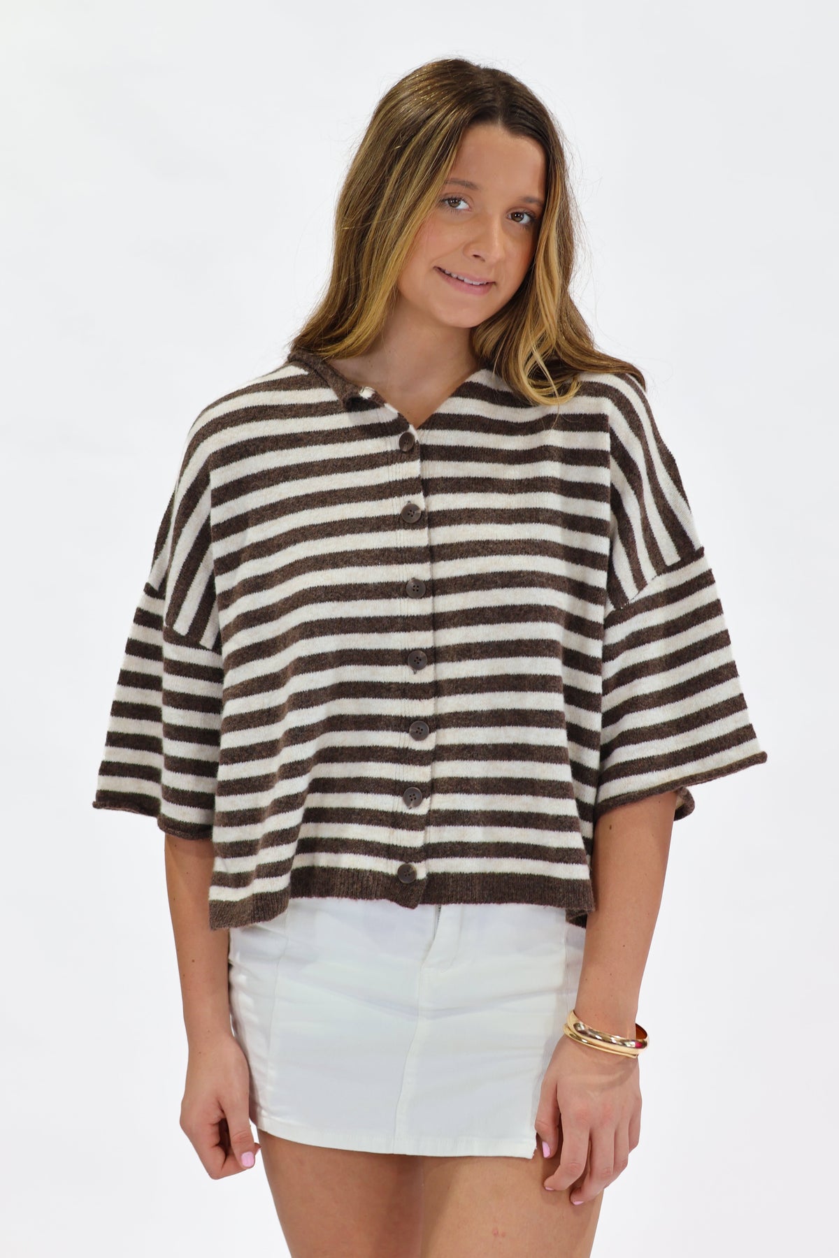 A woman modeling a brown and off-white horizontally striped short-sleeve button-up top with white shorts, set against a plain white studio background.