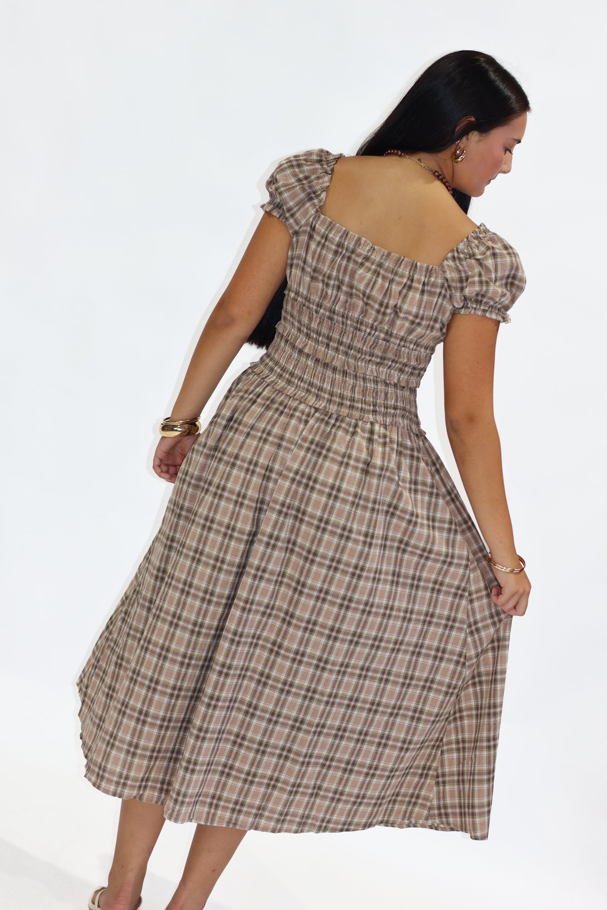A woman wearing a beige and brown plaid midi dress with puff sleeves and an off-the-shoulder neckline, shown from the back against a plain white studio background.