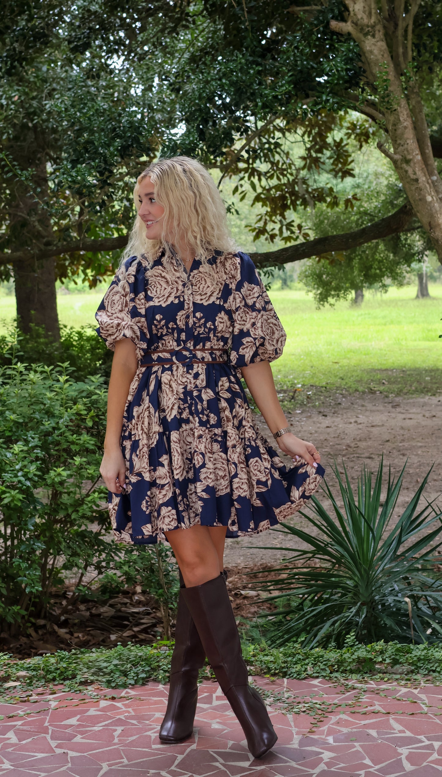 Woman in a floral dress and knee-high boots walking outdoors in a park-like setting.