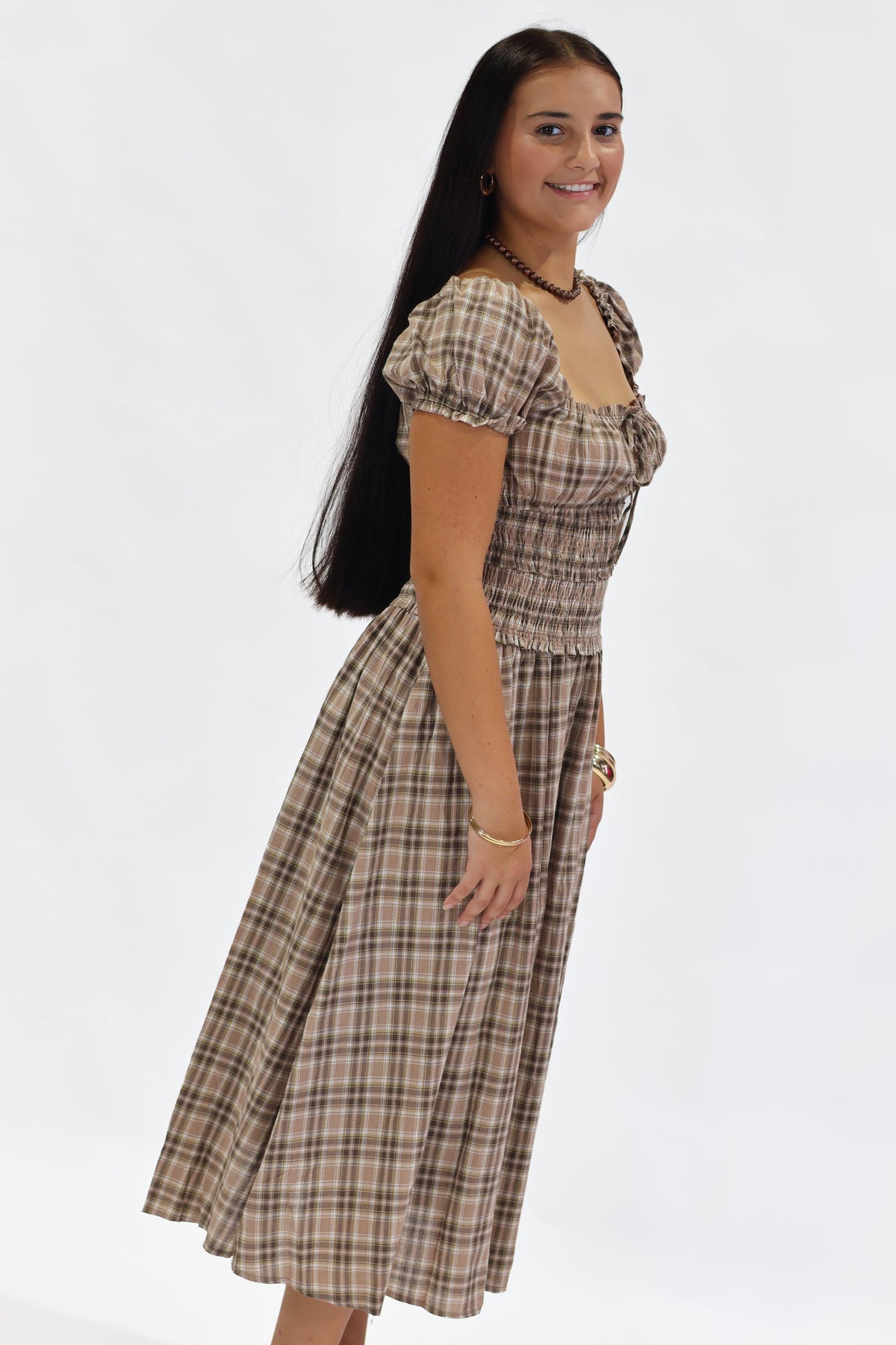 A woman modeling a brown and beige plaid midi dress with puff sleeves and a smocked bodice, standing against a plain white studio background.