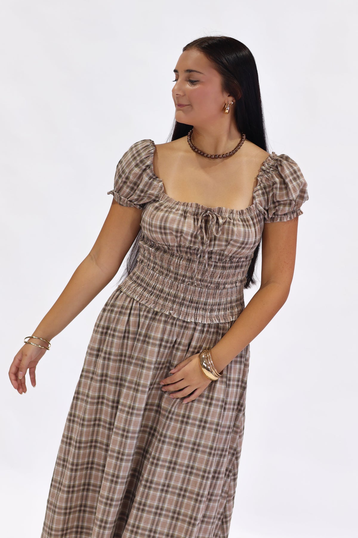 A woman modeling a brown and beige plaid midi dress with puff sleeves, a square neckline, and a smocked waist, set against a plain white studio background.