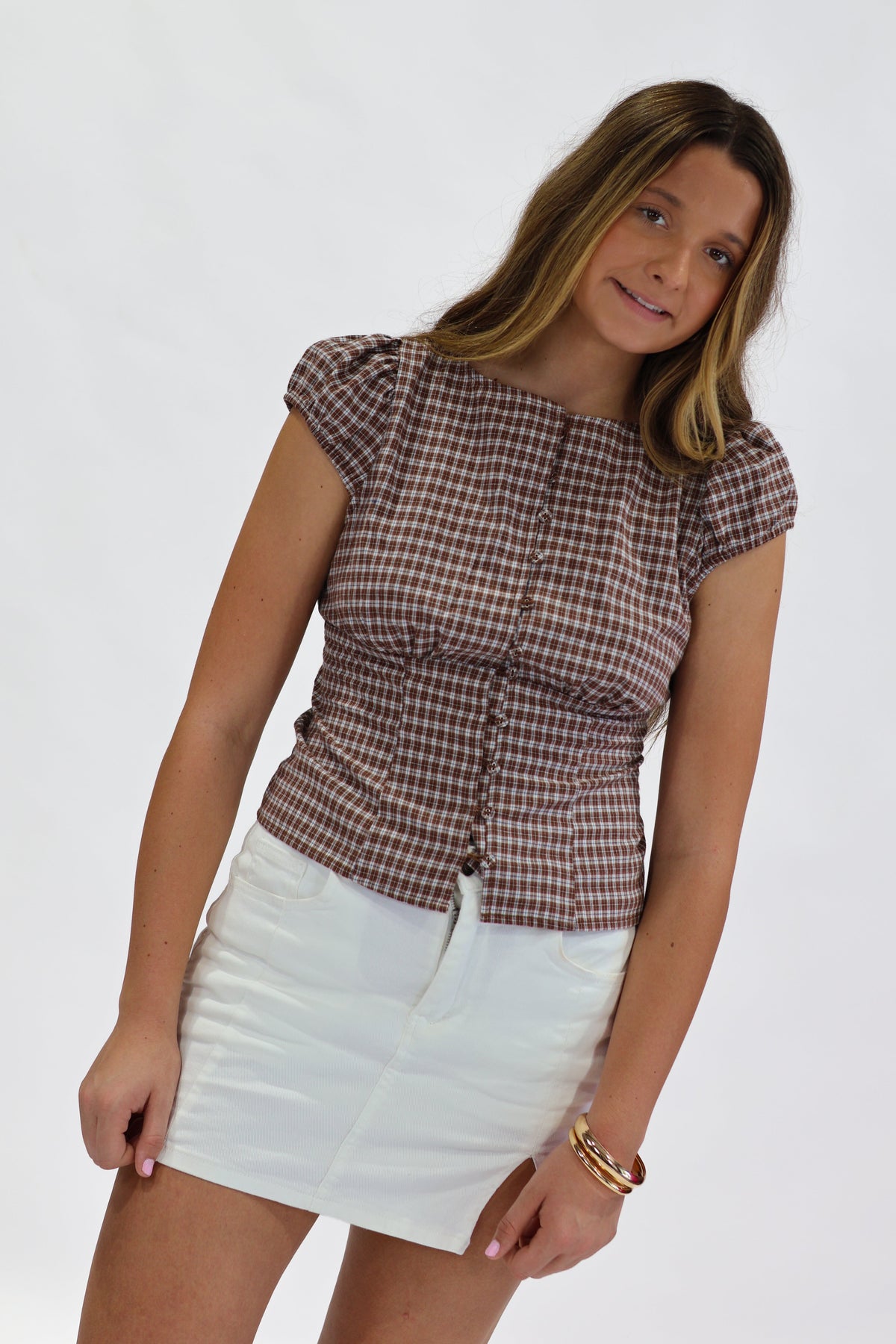 A woman modeling a brown and white checkered short-sleeve top with a gathered front, paired with a white mini skirt, against a plain white background.