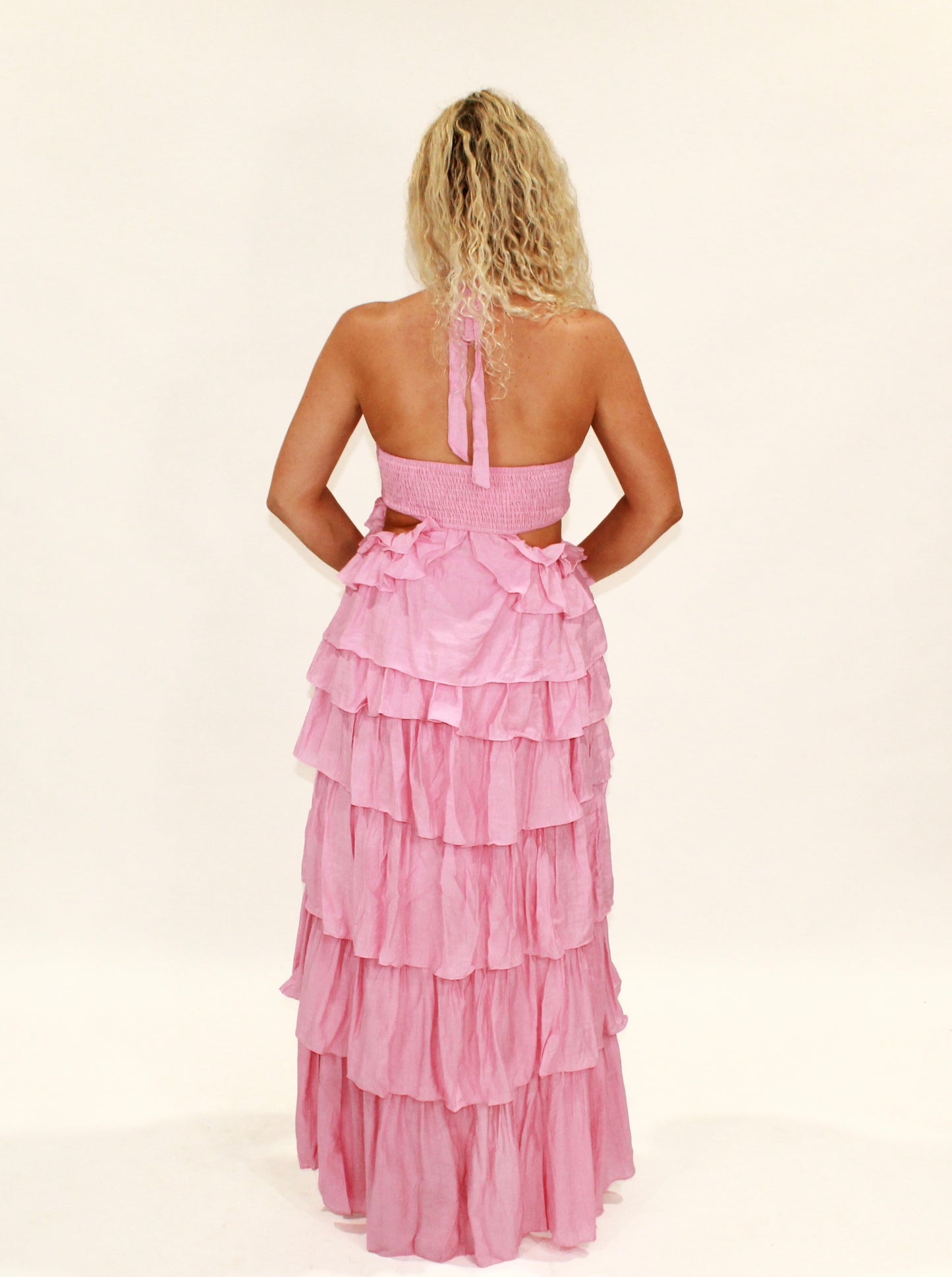 Back view of a tiered pink maxi dress, made of lightweight fabric, featuring a halter neck and ruffled details.
