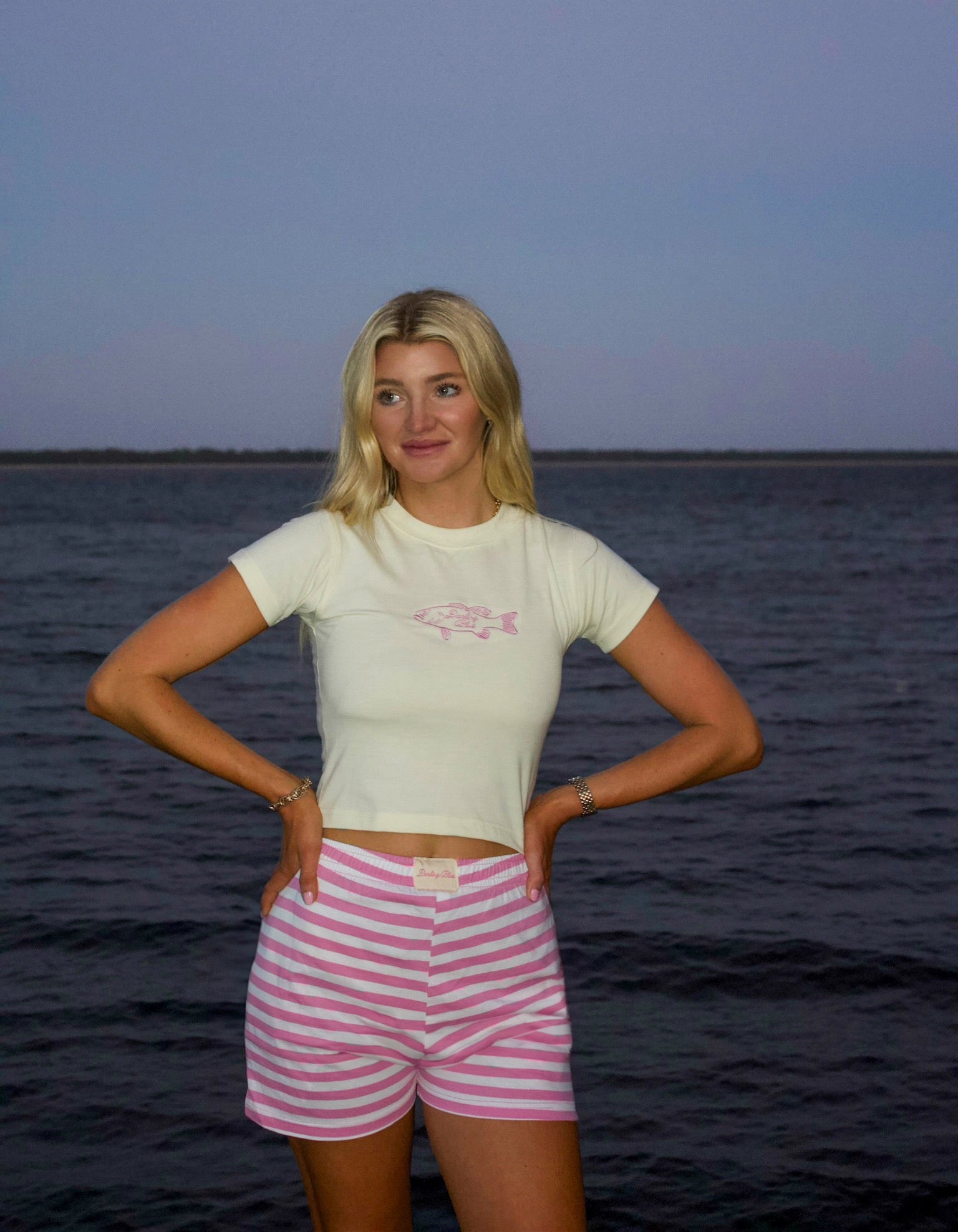 Woman standing by a body of water wearing a white shirt with a pink design and striped shorts.