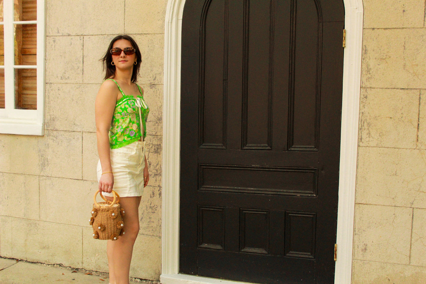 Woman in green patterned top and white skirt, holding a straw bag, standing by a black door.
