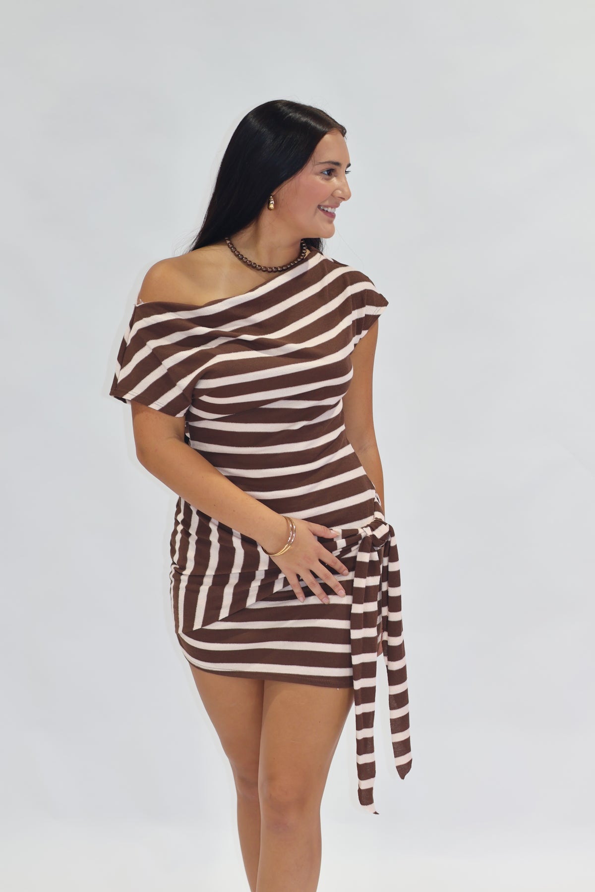 A woman modeling a brown and off-white horizontally striped off-the-shoulder mini dress with a side tie, set against a plain light gray studio background.
