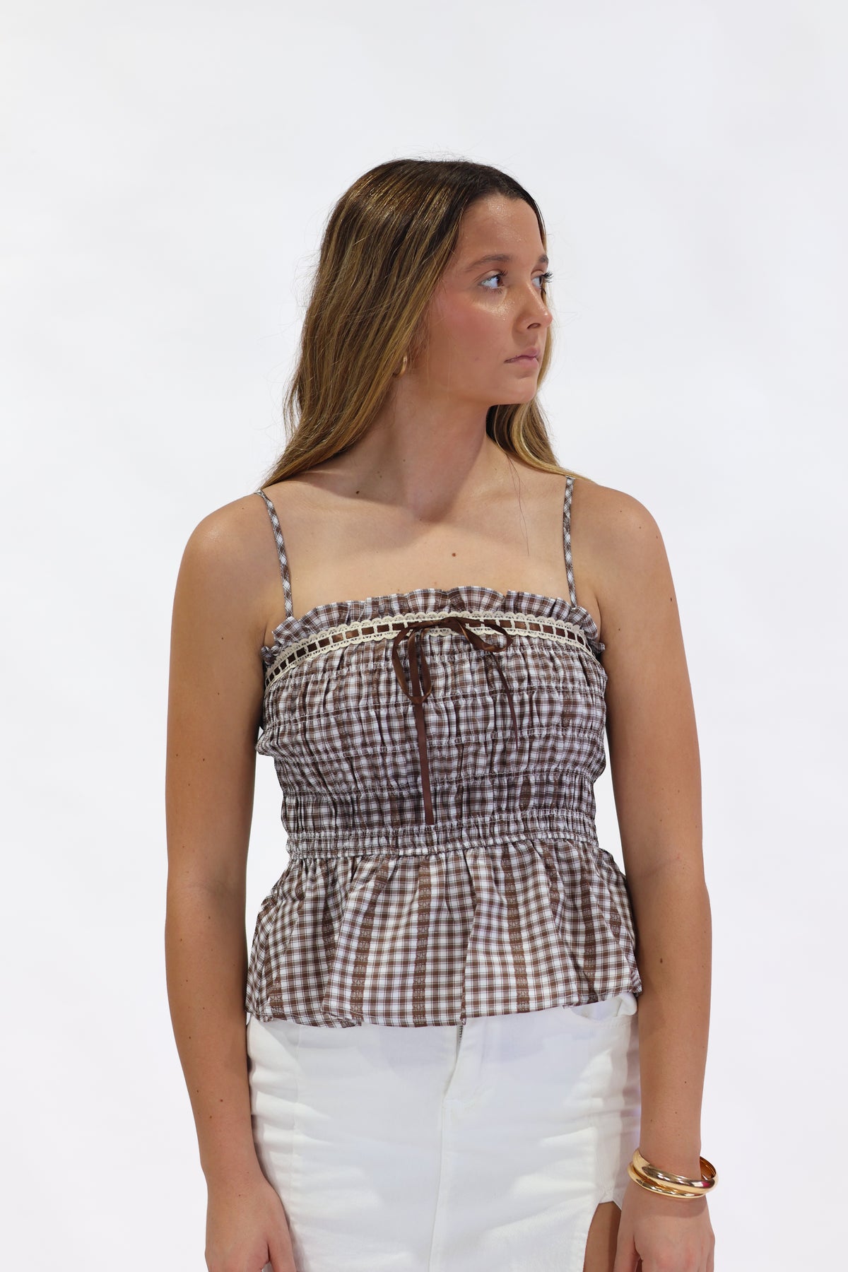 A woman modeling a brown and white gingham check strapless top with a smocked bodice, thin shoulder straps, and a ruffled peplum hem, paired with white pants against a plain white background.