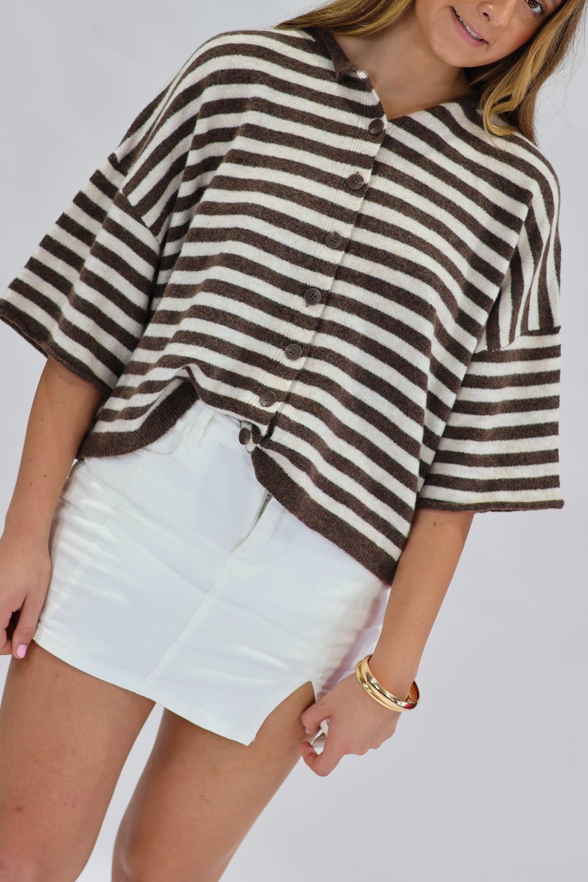 A woman modeling a brown and white horizontally striped cropped cardigan with wide sleeves, paired with a white mini skirt, against a light gray studio background.