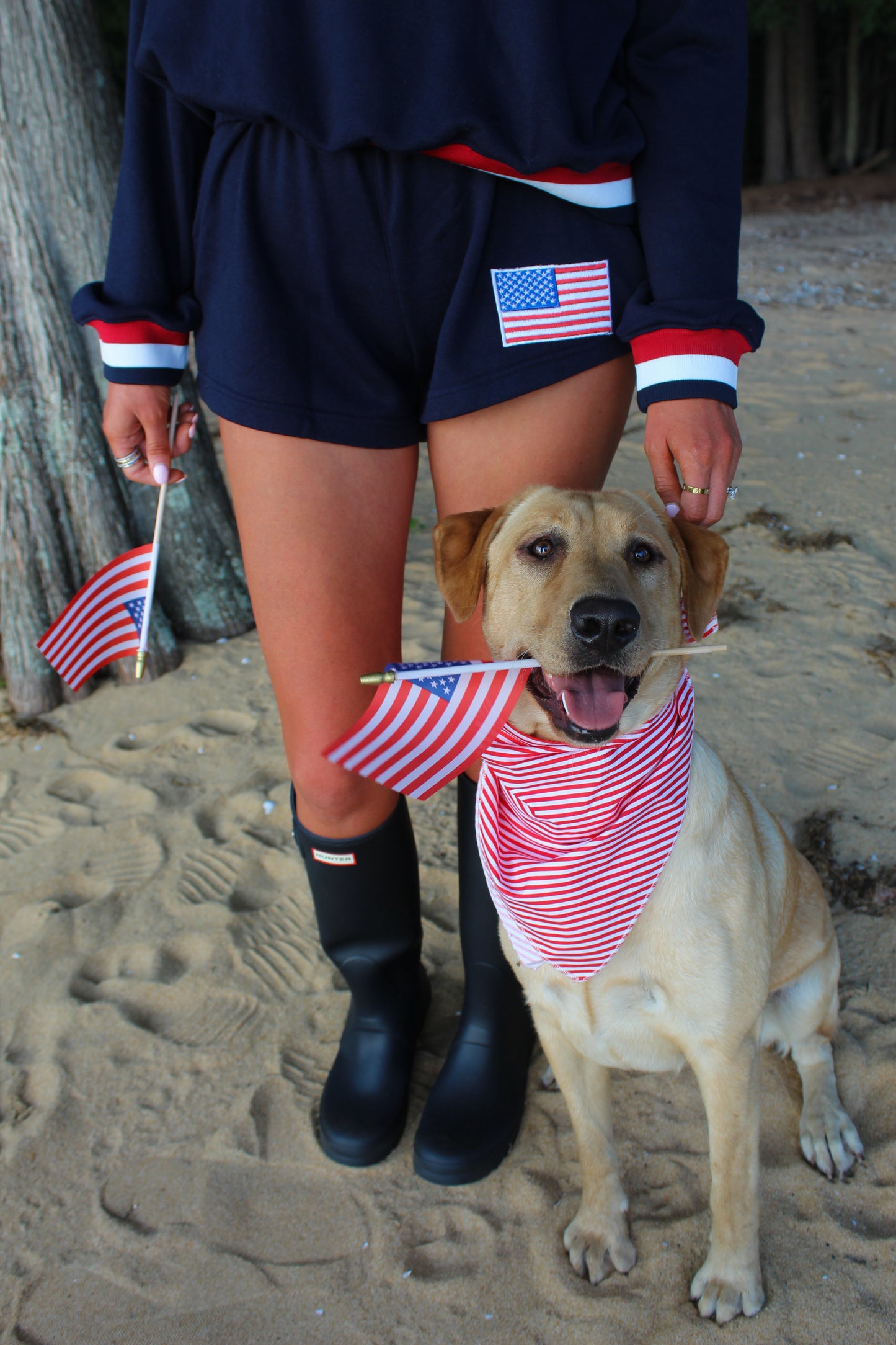 Woman in navy outfit with red accents holding American flags next to a happy dog in a striped bandana.
