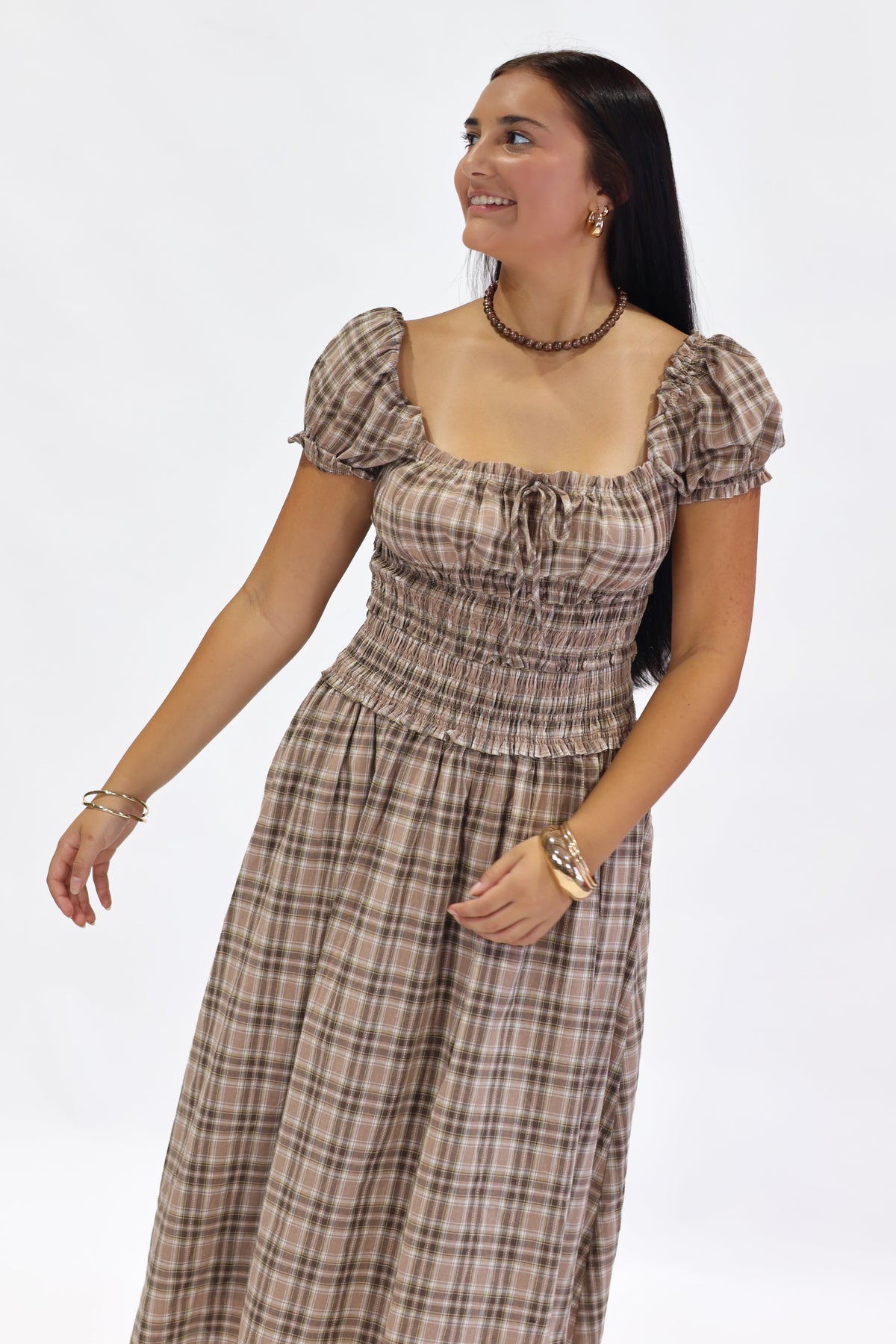 A woman modeling a brown and beige plaid midi dress with puff sleeves, a smocked bodice, and a tie at the neckline, set against a plain white studio background.