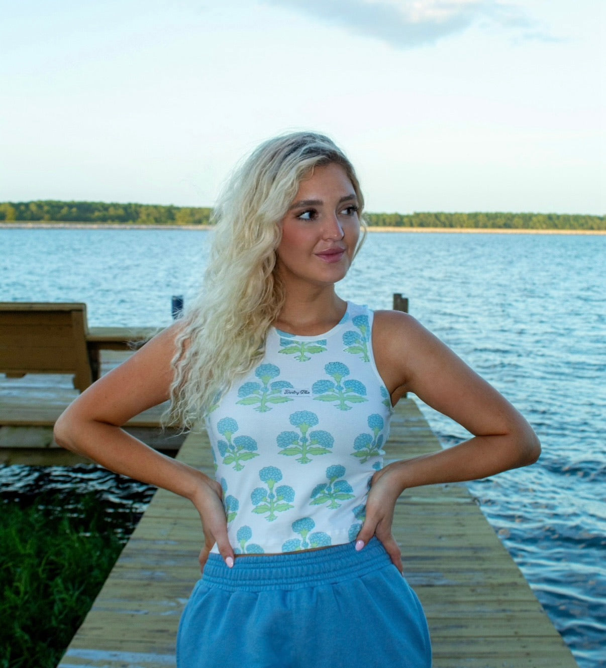 Woman standing on a dock by a lake wearing a patterned top and blue pants.
