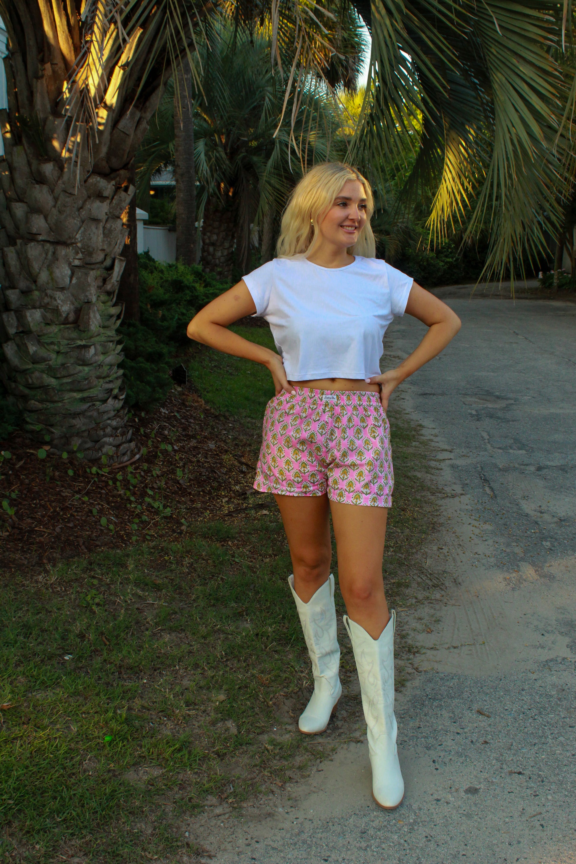 Woman in a white crop top and pink patterned shorts, wearing white cowboy boots in a tropical setting.