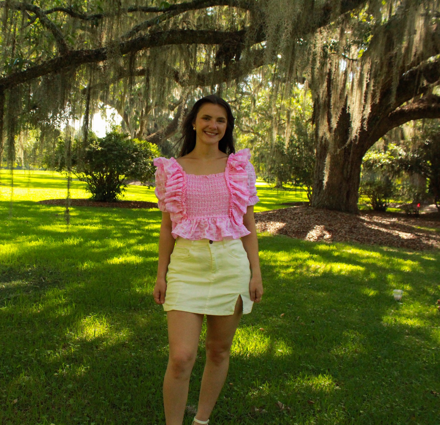 Model wearing a pink ruffled smocked top and white skirt in a sunny outdoor setting.