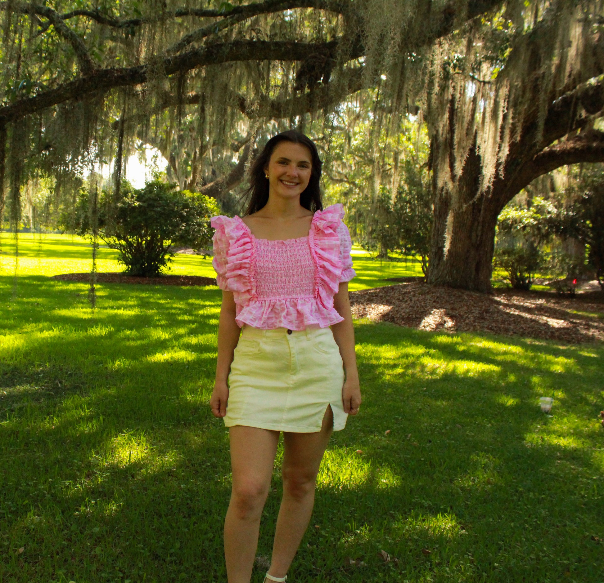 Model wearing a pink ruffled smocked top and white skirt in a sunny outdoor setting.