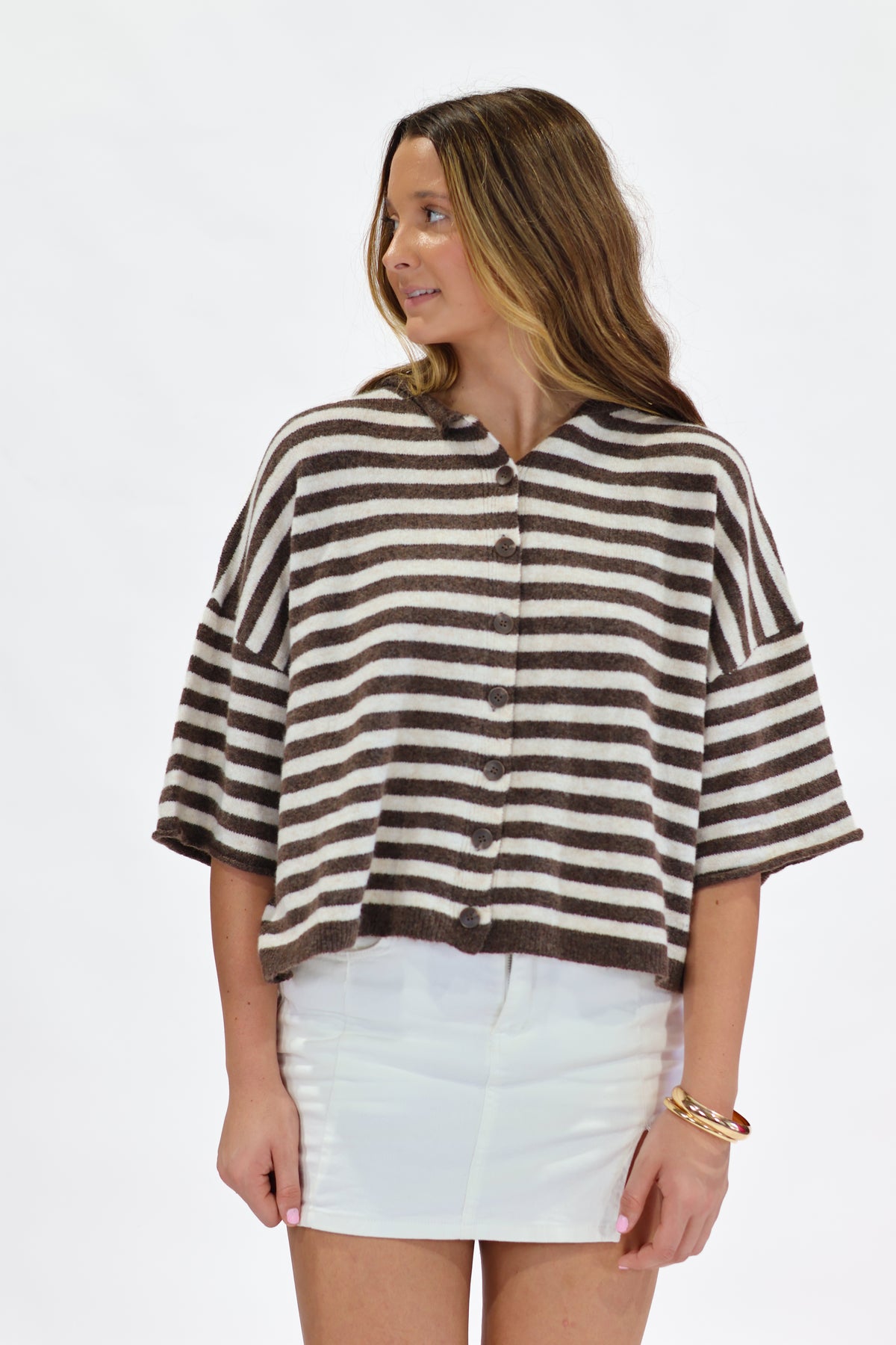 A woman wearing a brown and cream striped short-sleeve button-up top with white shorts, standing against a plain white studio background.