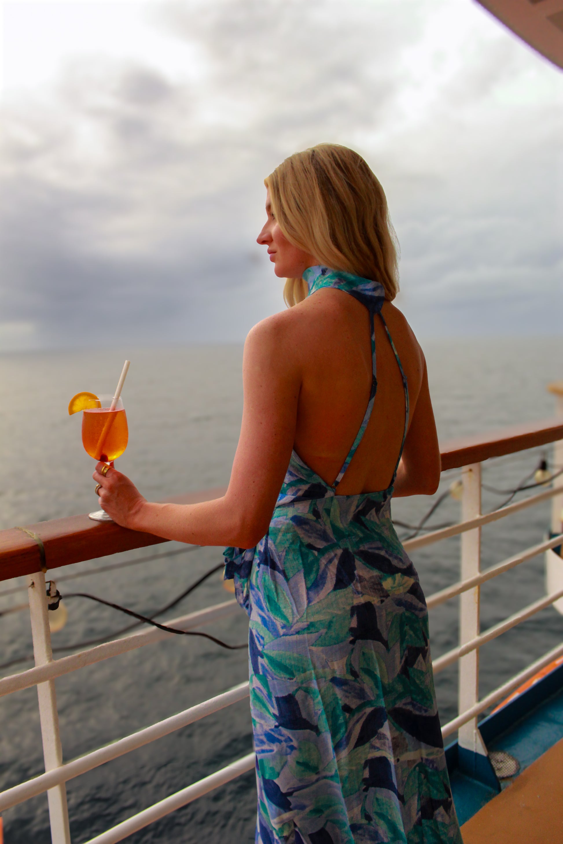 Woman in a blue tropical print halter dress enjoying a drink on a cruise ship balcony.