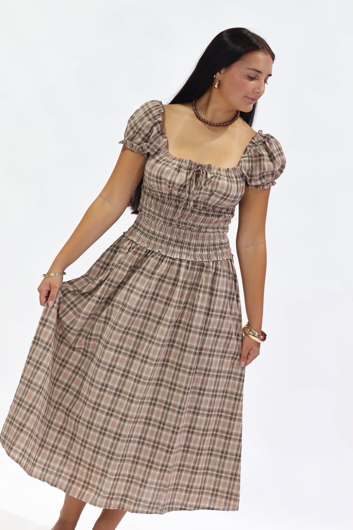A woman modeling a brown and beige plaid midi dress with puff sleeves, a square neckline, and a smocked waist, set against a plain white studio background.