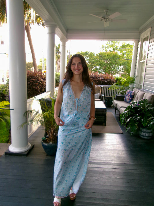 Woman in a light blue floral maxi dress with a v-neckline, walking on a porch surrounded by greenery.