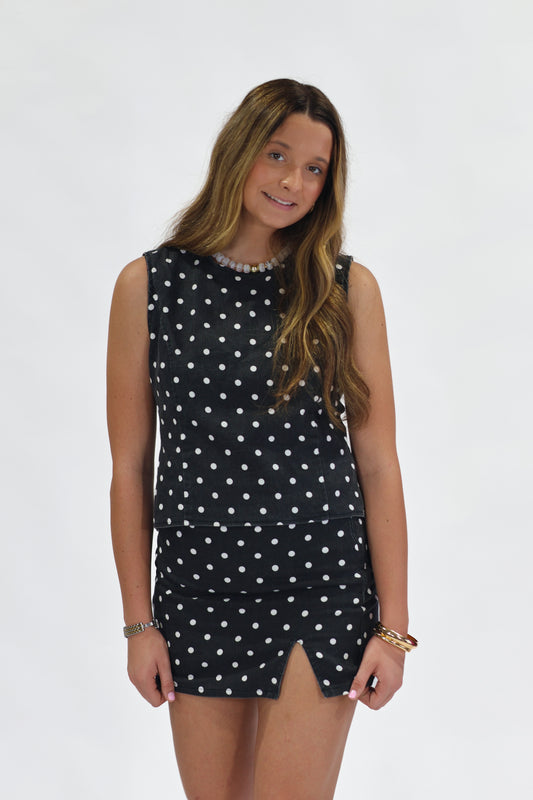 A woman modeling a sleeveless black dress with a white polka dot pattern, standing against a plain white studio background.