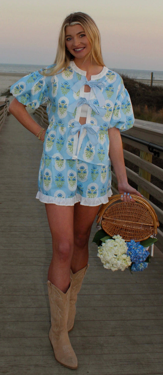 Floral women's romper in blue, featuring puff sleeves and lace trim, styled with cowboy boots and a basket.
